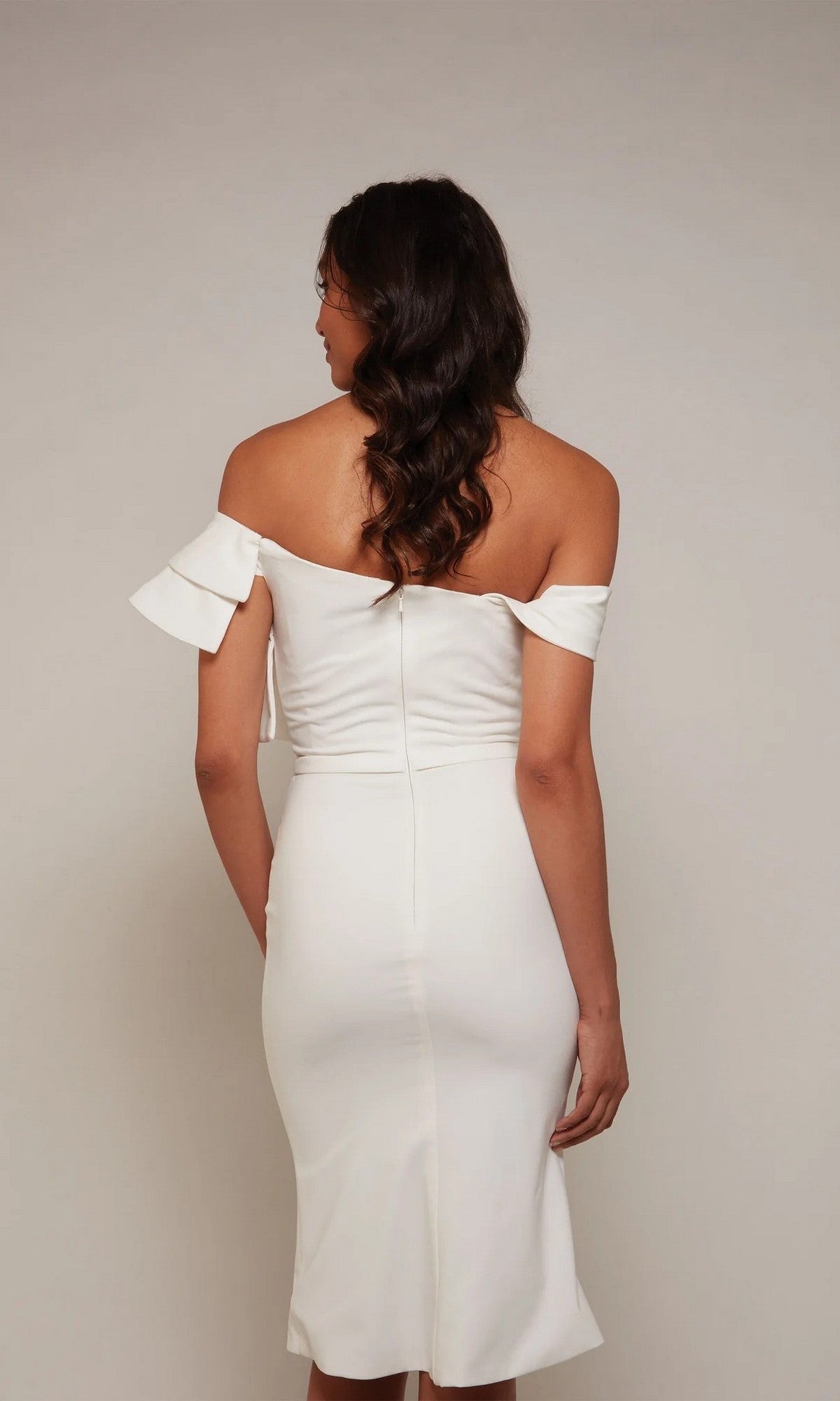 A woman faces away, wearing the Alyce 70032 Knee Length White Party Dress—a fitted off-the-shoulder midi with ruffle sleeves—her wavy hair cascading down her back.